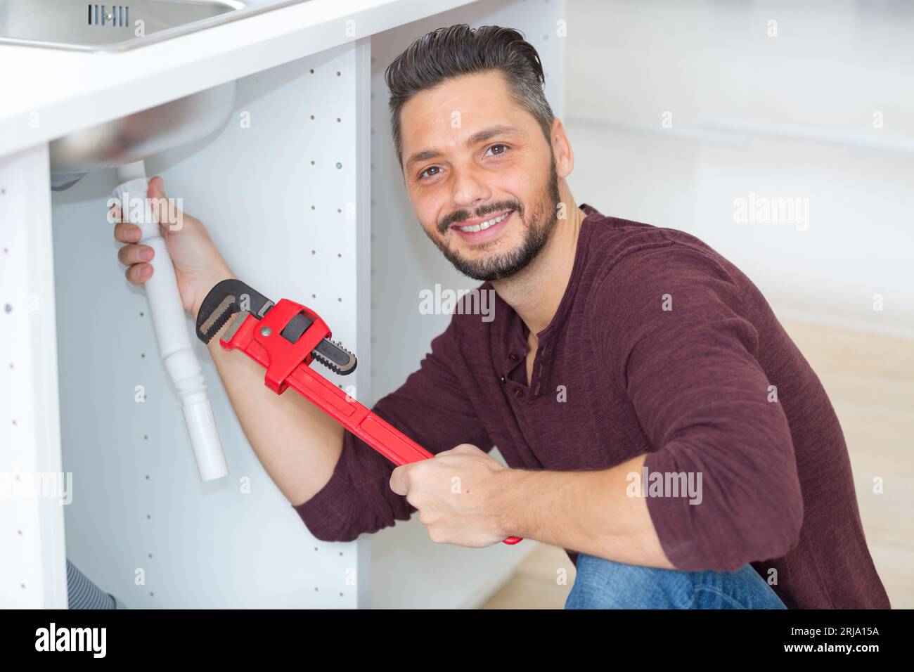 man fitting sink drainage pipe under kitchen sink holding wrench Stock