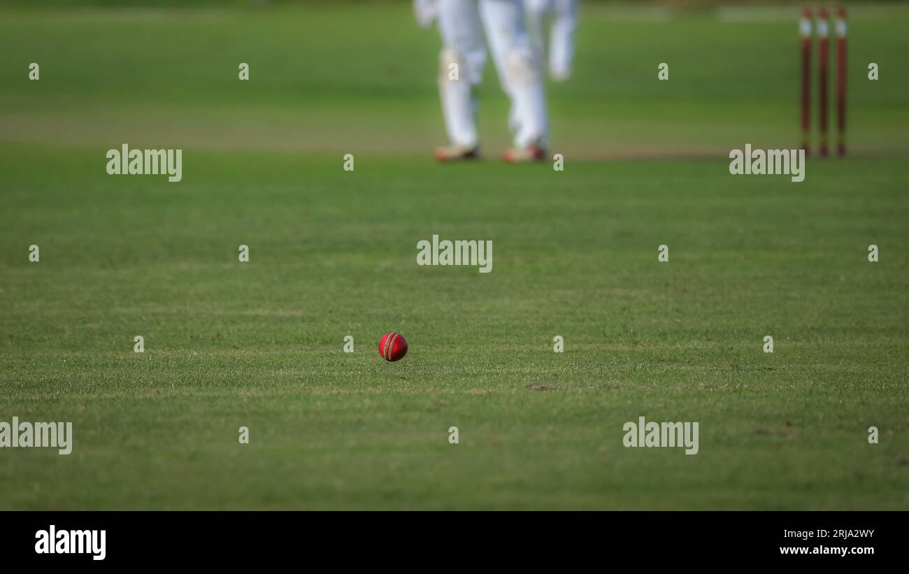 A red cricket ball, inches above the grass and heading for the boundary.  The stumps and part of a player are visible in the out of focus background Stock Photo