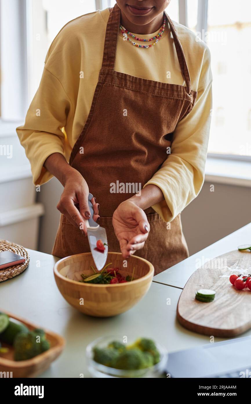 Vertical portrait of young black woman adding vegetables to wooden bowl ...