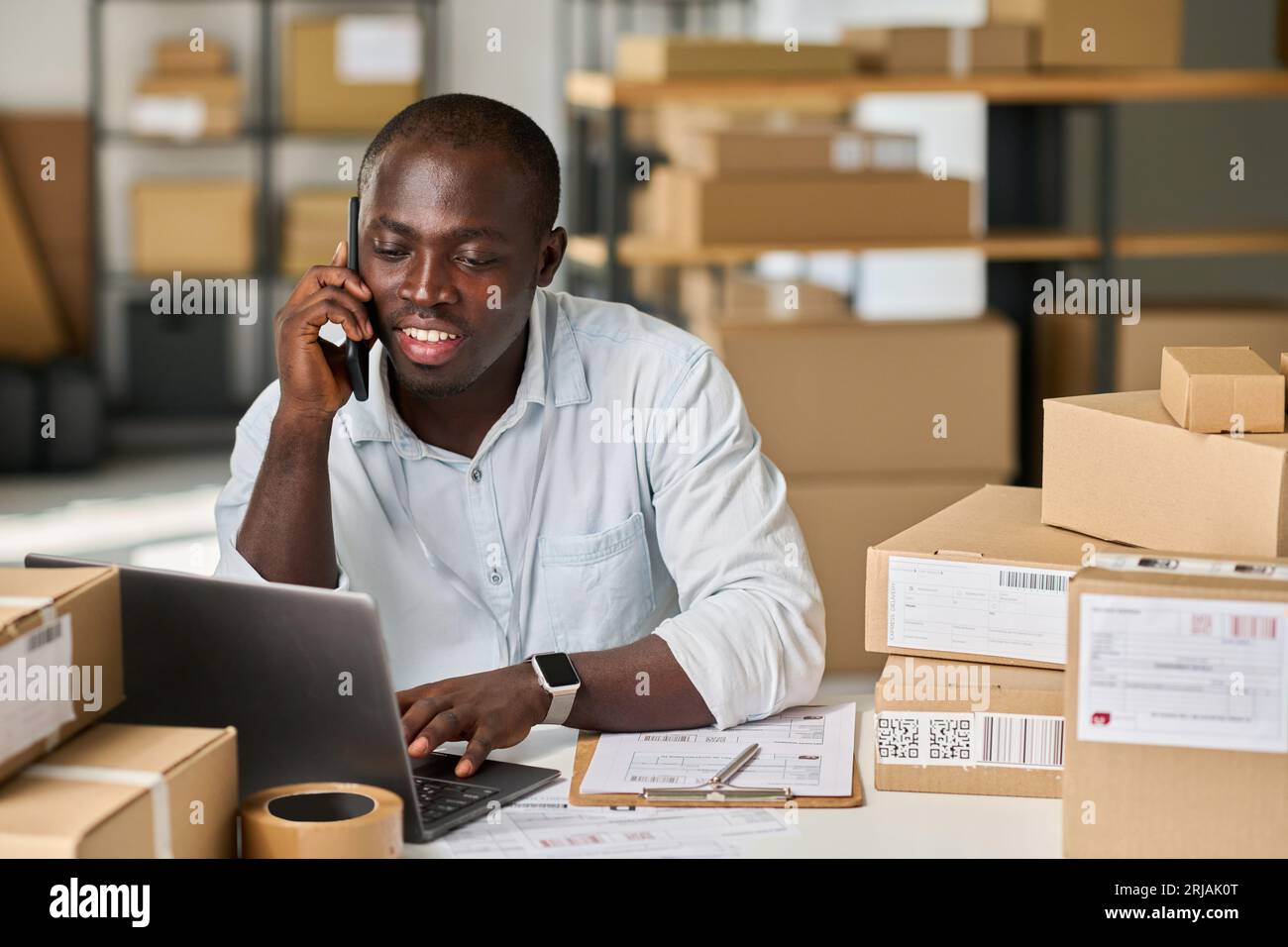 Young African American man in white shirt sitting by workplace in warehouse or marketplace and talking on mobile phone to client Stock Photo