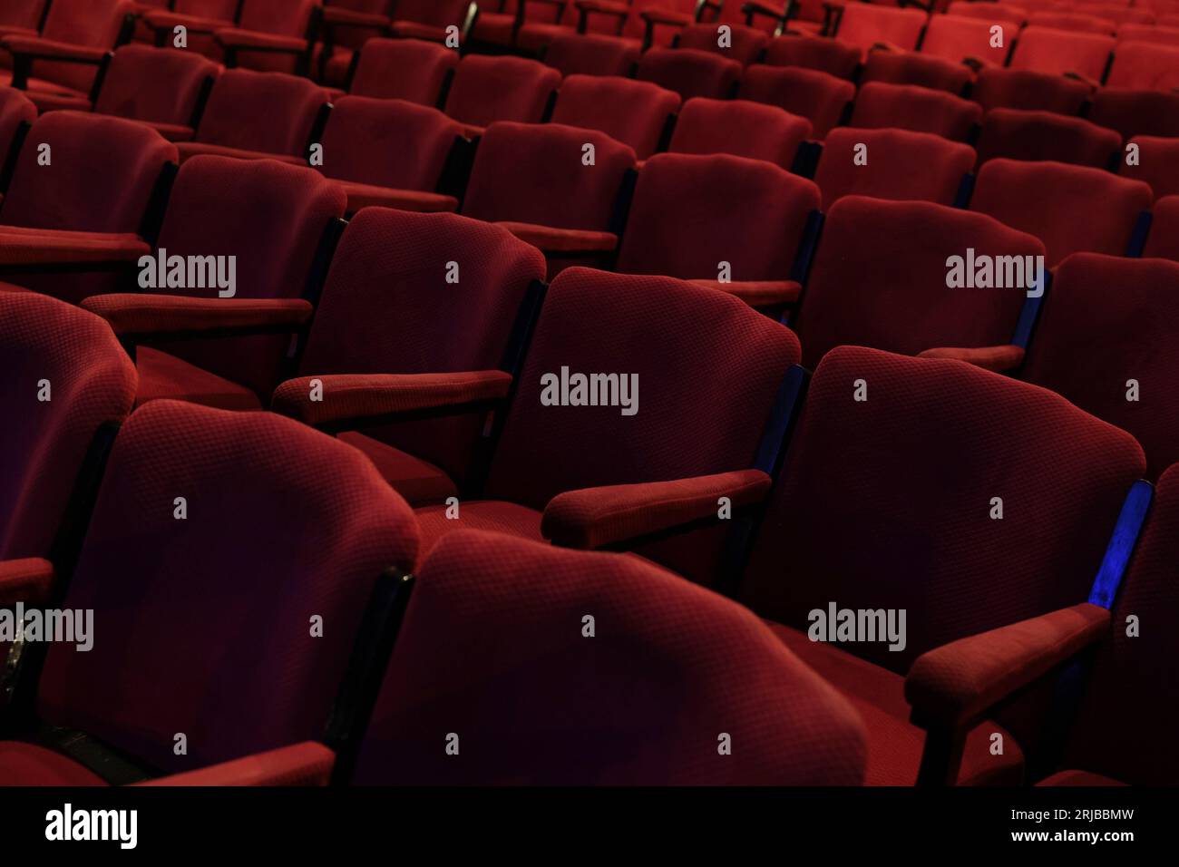 Empty rows of deep red fabric chairs or seats with arm rests in a theater or movie cinema hall. Stock Photo