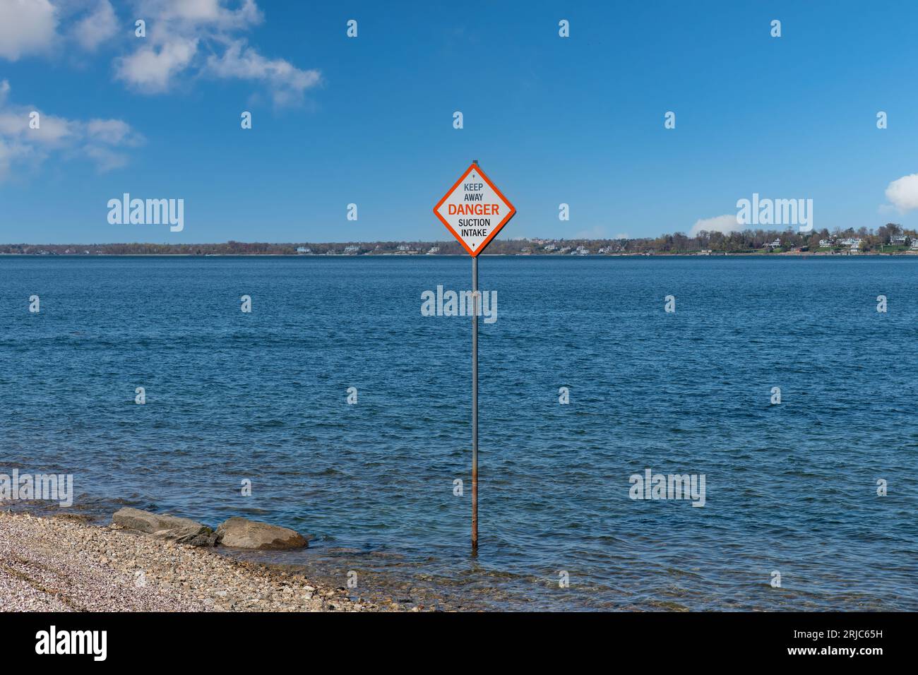 View over a large body of water with in front on shore a warning sign on a post advising of danger and to keep away from the suction intake Stock Photo