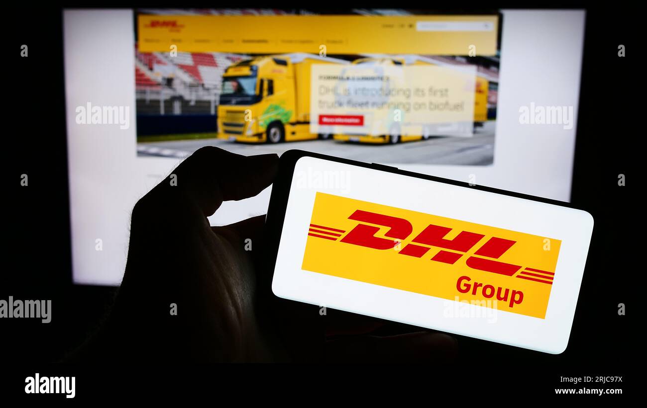 Person holding cellphone with logo of logistics company Deutsche Post AG (DHL Group) on screen in front of webpage. Focus on phone display. Stock Photo