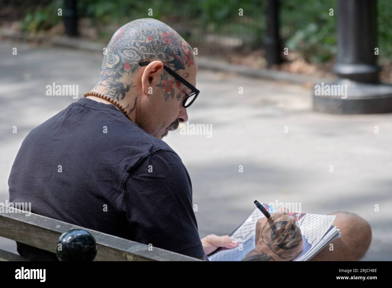 A middle aged man with a fully tattooed shaved head draws while sitting on a bench in Washington Square Park in Greenwich Village. Stock Photo