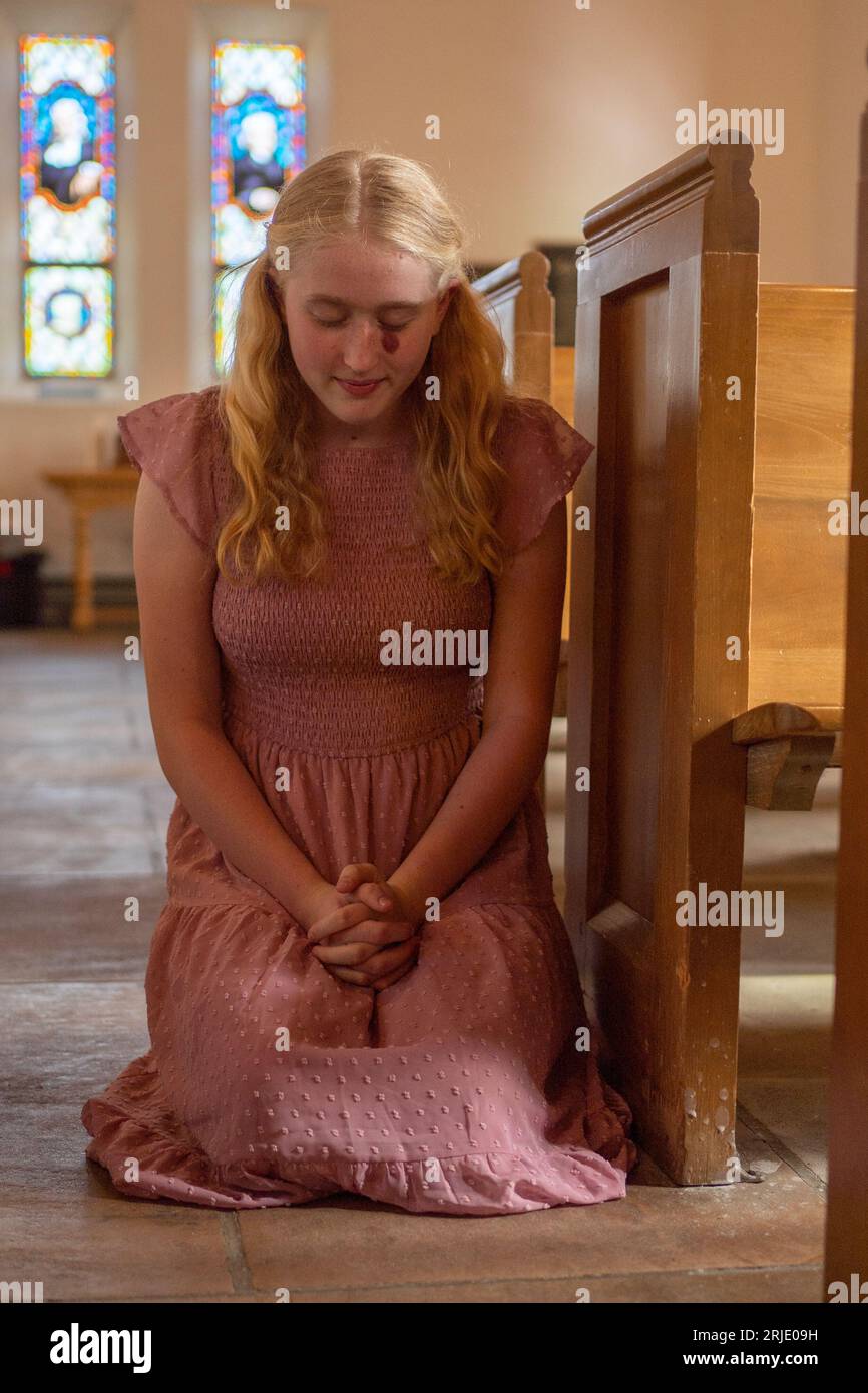 A young female kneeling in prayer inside a church, her hands clasped ...