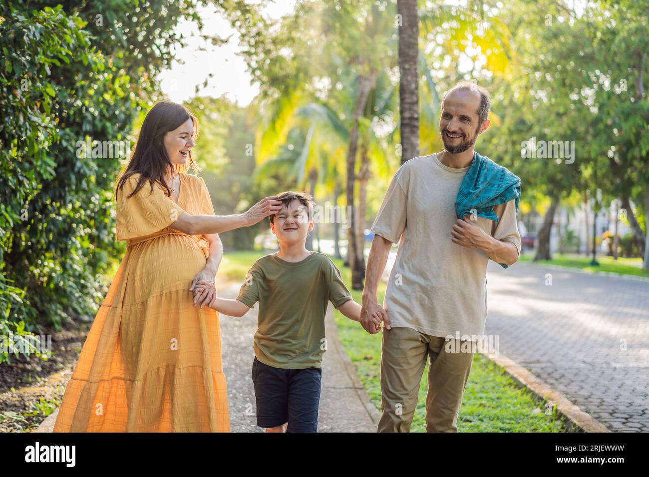 A loving family enjoying a leisurely walk in the park - a radiant pregnant woman after 40, embraced by her husband, and accompanied by their adult Stock Photo