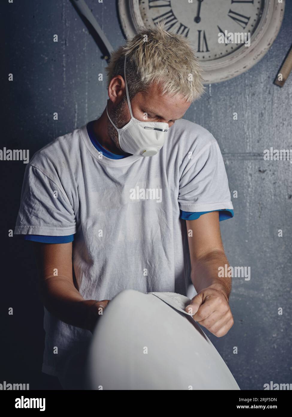 Focused adult blond haired tattooed male master grinding white handmade surfboard while shaping it with sandpaper in workshop Stock Photo