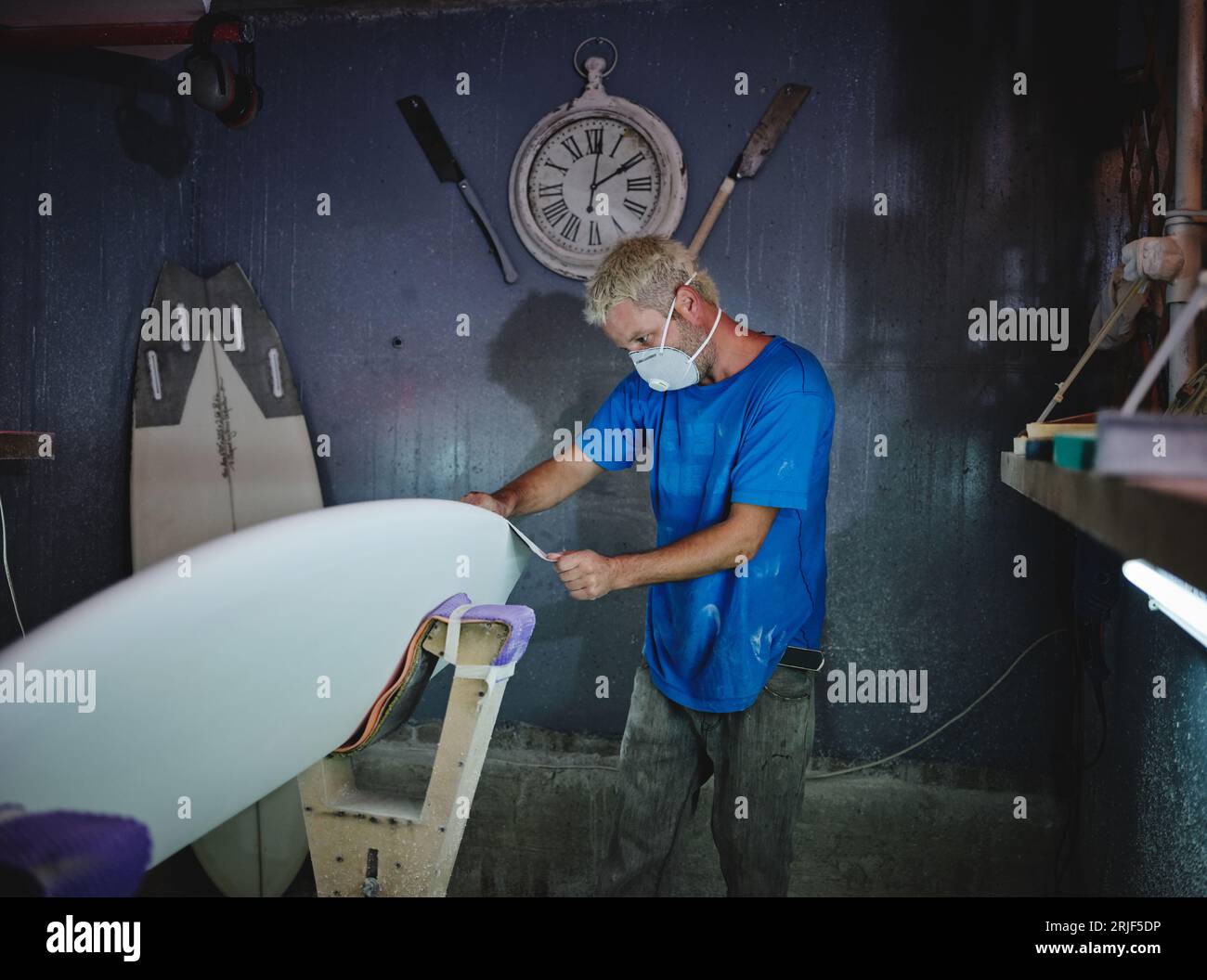 Side view of adult blond haired master in respirator mask grinding white surfboard placed in shaping racks while working in workshop Stock Photo