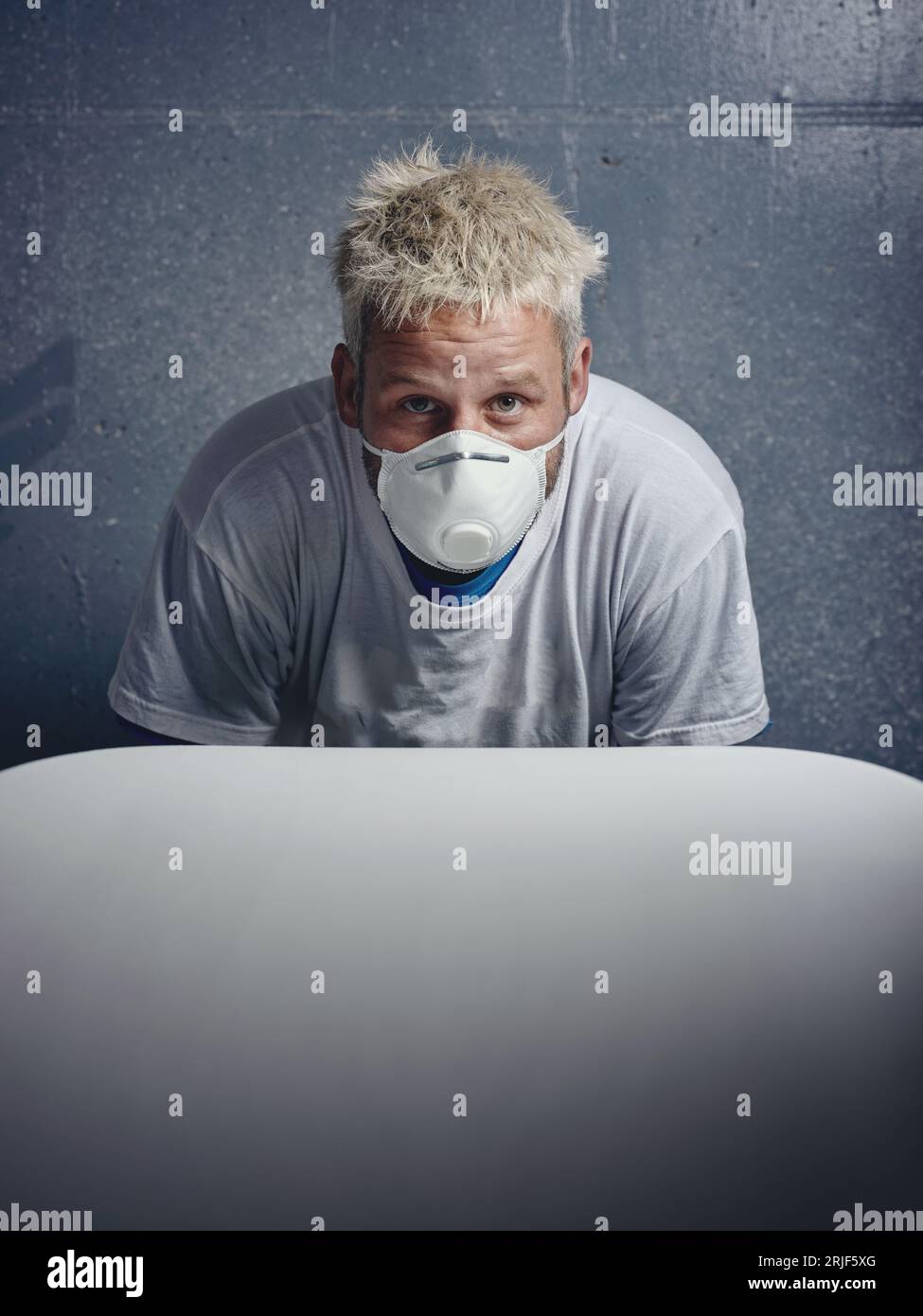 Focused blond haired male shaper wearing protective respirator mask while making white surfboard and looking at camera against gray wall Stock Photo
