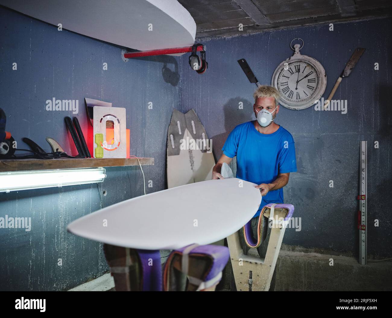 Concentrated blond haired male shaper in respirator mask examining shape of board placed on racks while creating handmade surfboards in workshop Stock Photo