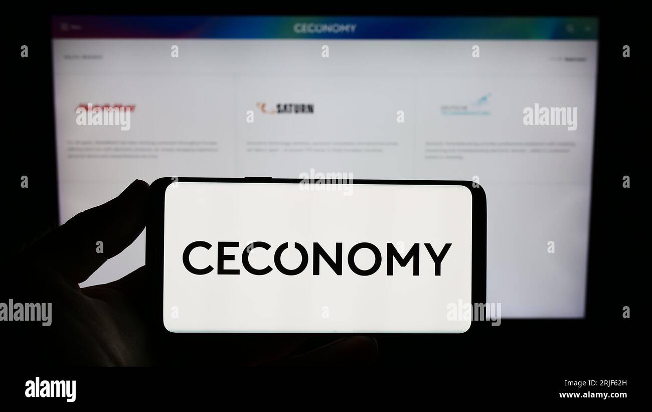 Person holding smartphone with logo of German retail company Ceconomy AG on screen in front of website. Focus on phone display. Stock Photo