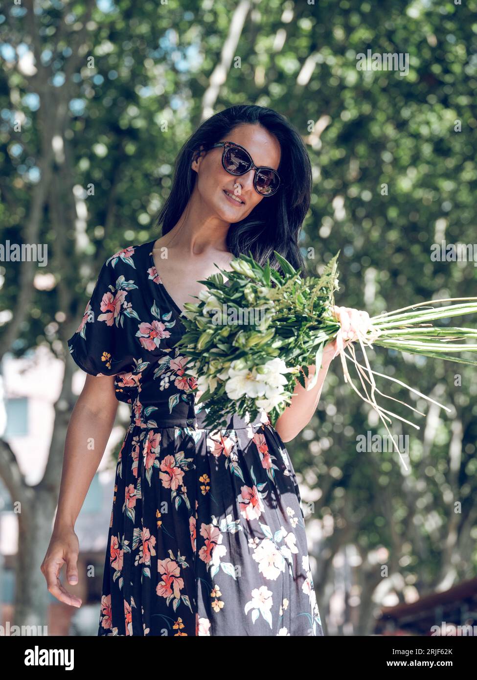 Charming smiling female with dark hair wearing summer dress and sunglasses holding bouquet of fresh white Alstroemeria flowers while walking in sunny Stock Photo