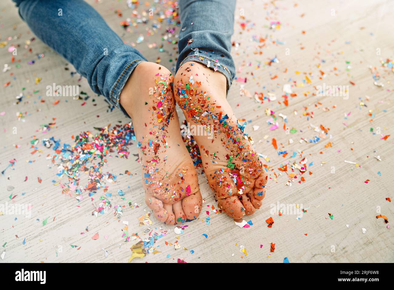 Top view of crop unrecognizable barefoot child in jeans lying on wooden light floor covered with colorful confetti Stock Photo
