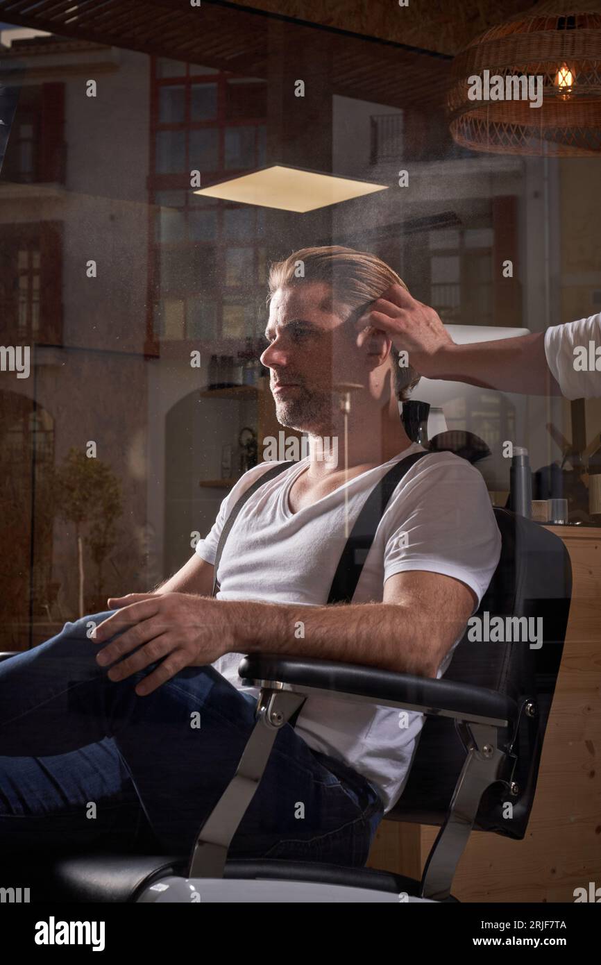 Confident male hipster sitting in armchair and getting new haircut by professional hairstylist in barbershop Stock Photo