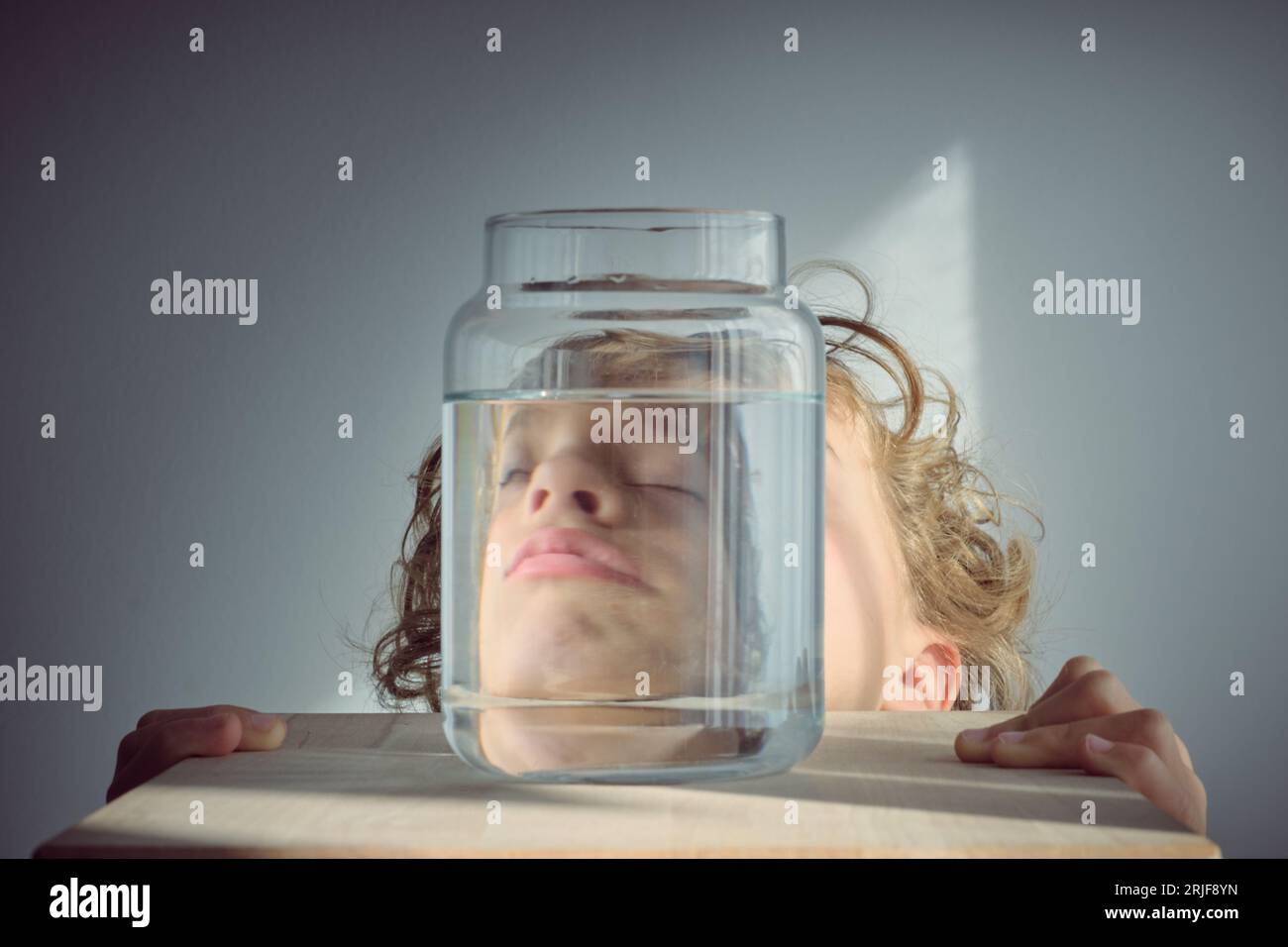 Sleepy curly blond haired kid reflection in glass transparent bottle of clear water against white background Stock Photo