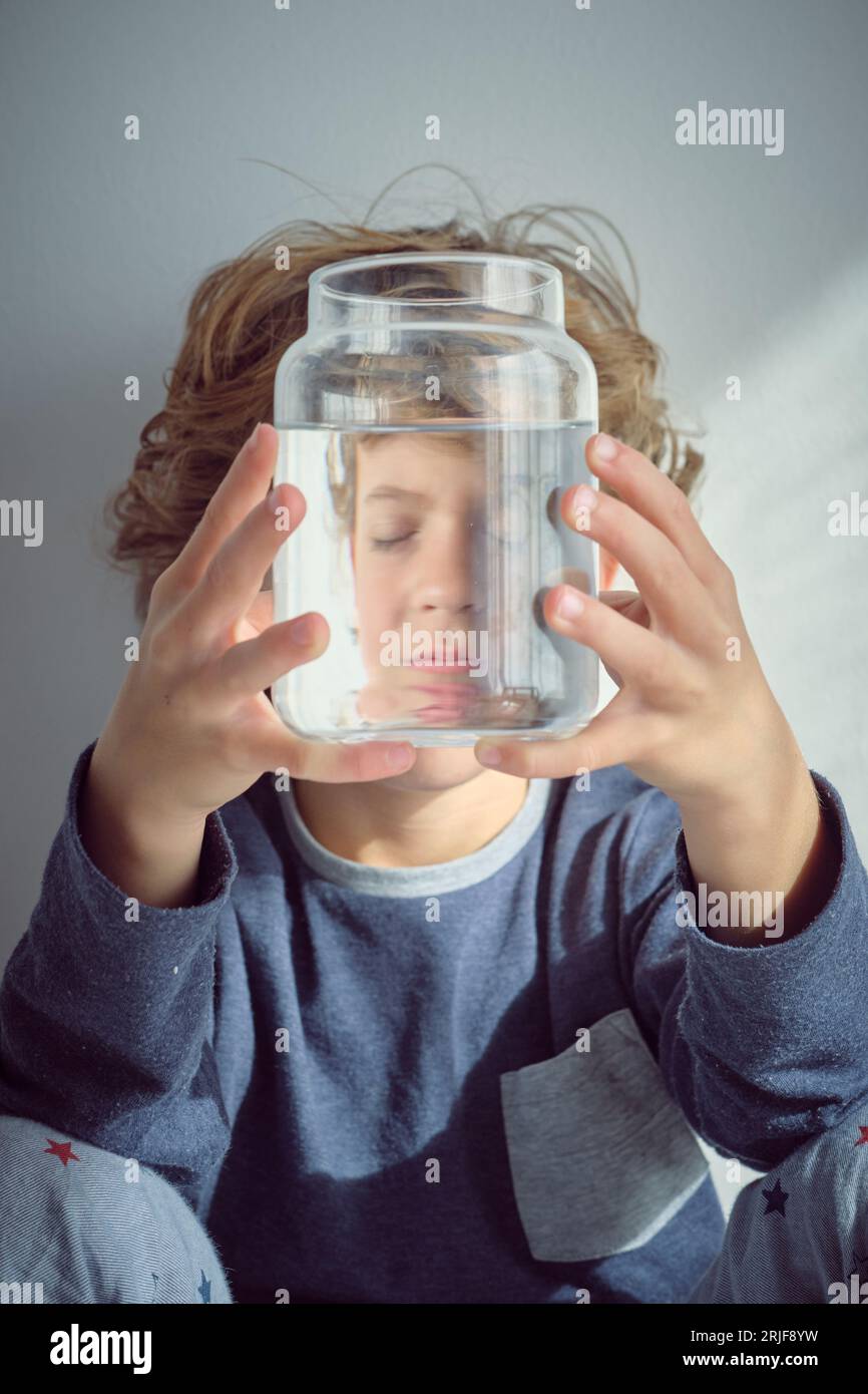 Blond haired kid with closed eyes covering face with glass jar of clean water white siting near white wall Stock Photo