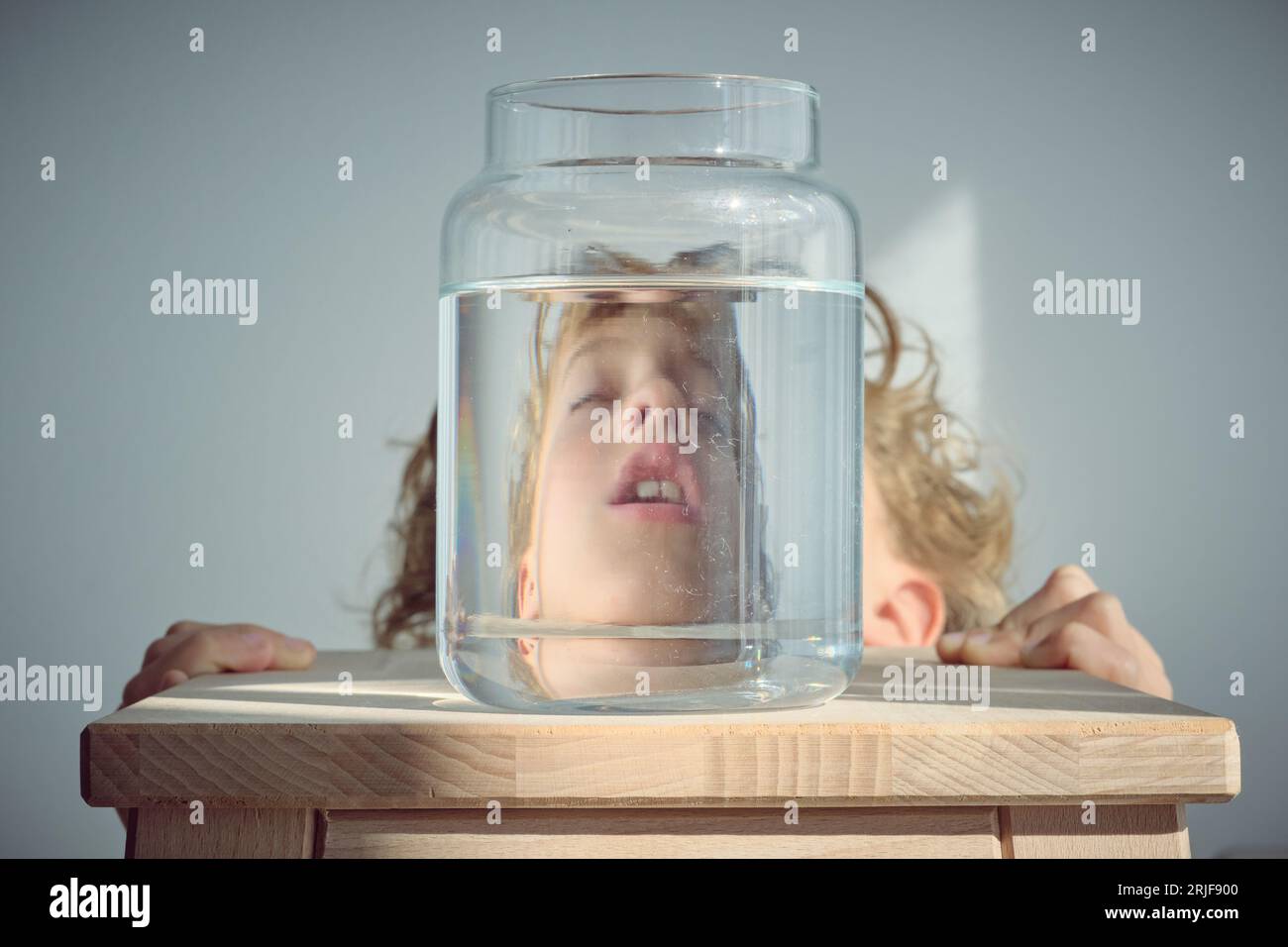 Napping kid with blond hair and open mouth near glass jar of water placed on wooden stool against white background Stock Photo