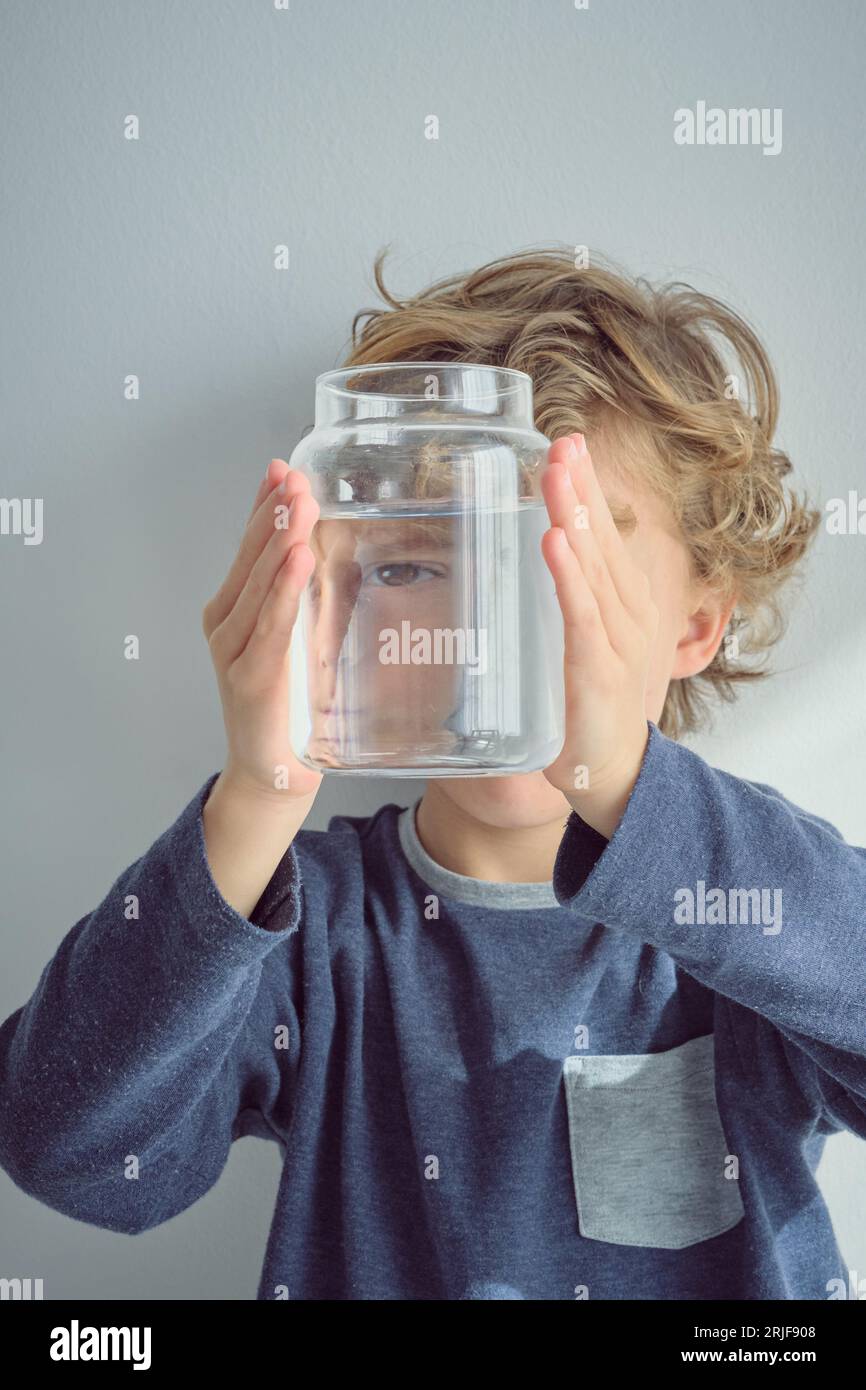 Blond haired boy looking through transparent glass bottle of water while holding jar against face near white wall Stock Photo