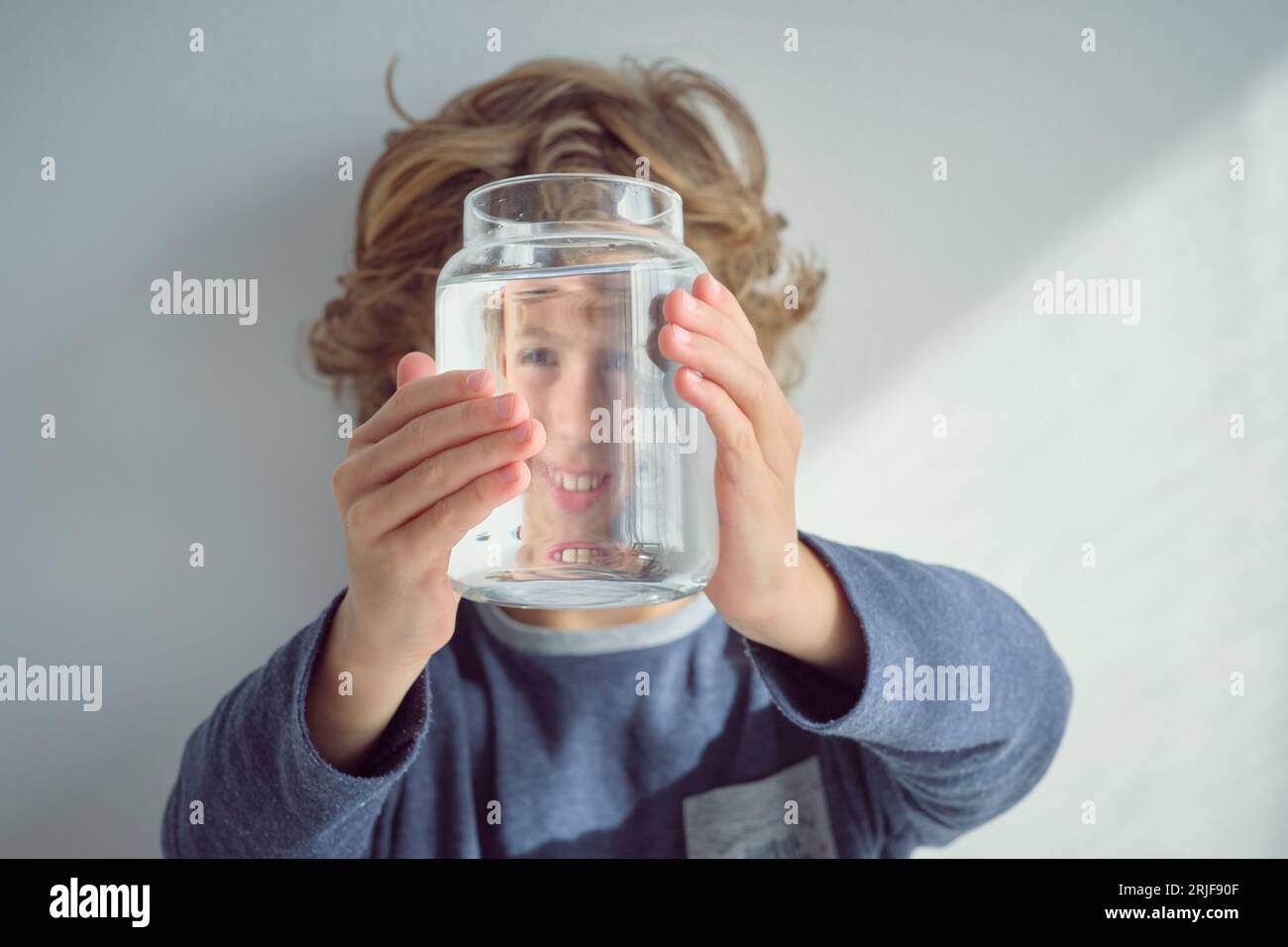 Cheerful blond haired kid looking through glass transparent bottle filled with clean water and holding jar against face near white background Stock Photo