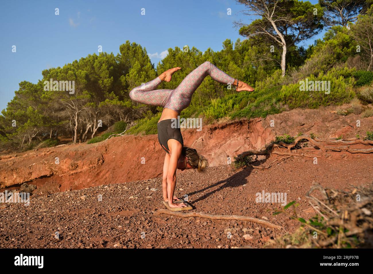 Woman in tights practicing a variation of handstand yoga pose with her feet up and her hands resting on a wooden board on a path in the middle of the Stock Photo