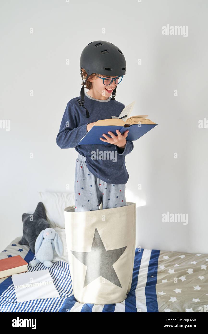 Concentrated preteen child in eyeglasses and helmet smiling while standing on bed at home and reading interesting book Stock Photo