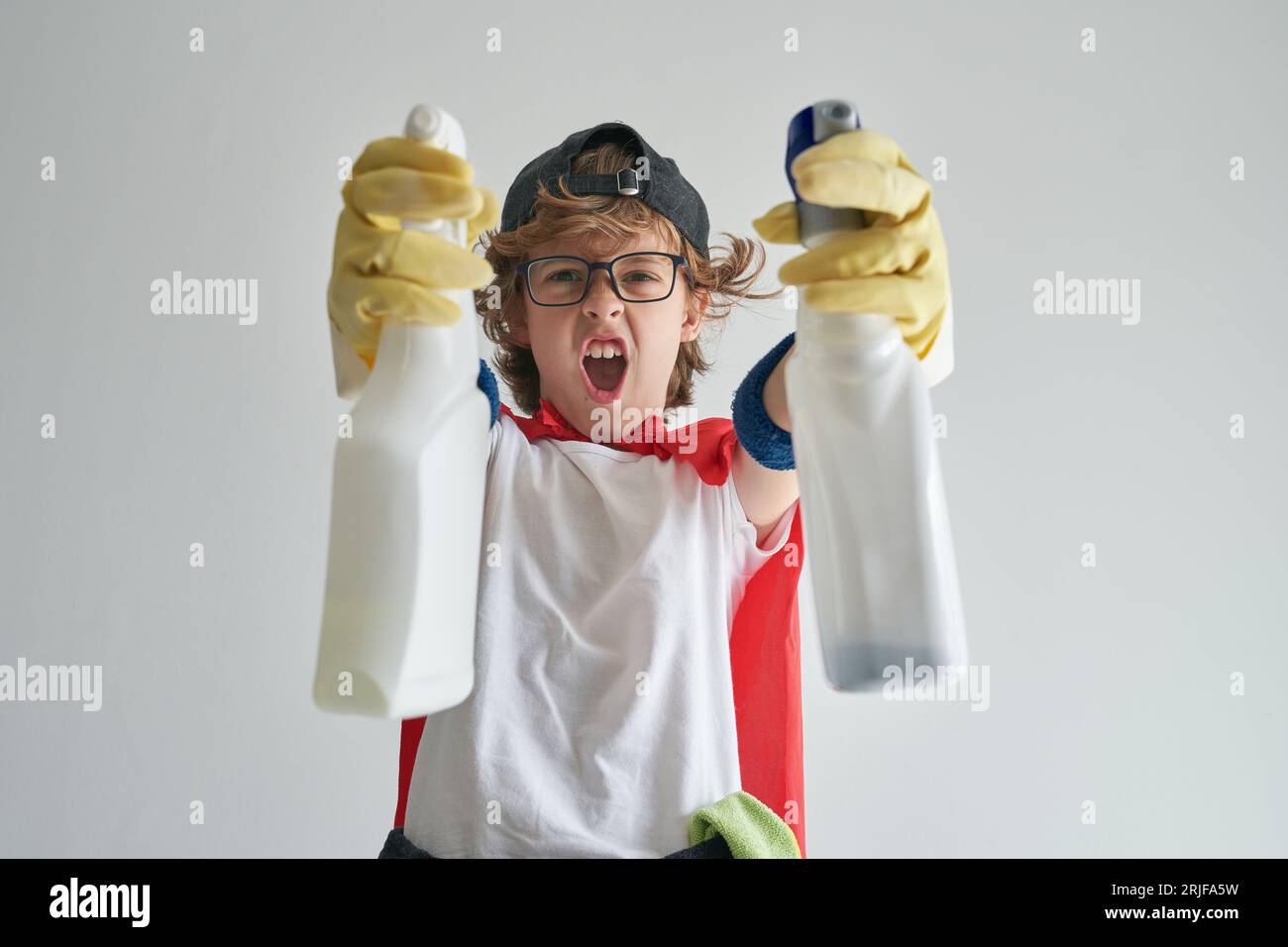 Funny preteen boy showing white plastic detergent bottles with hands in yellow gloves while looking at camera and screaming loudly with opened mouth d Stock Photo