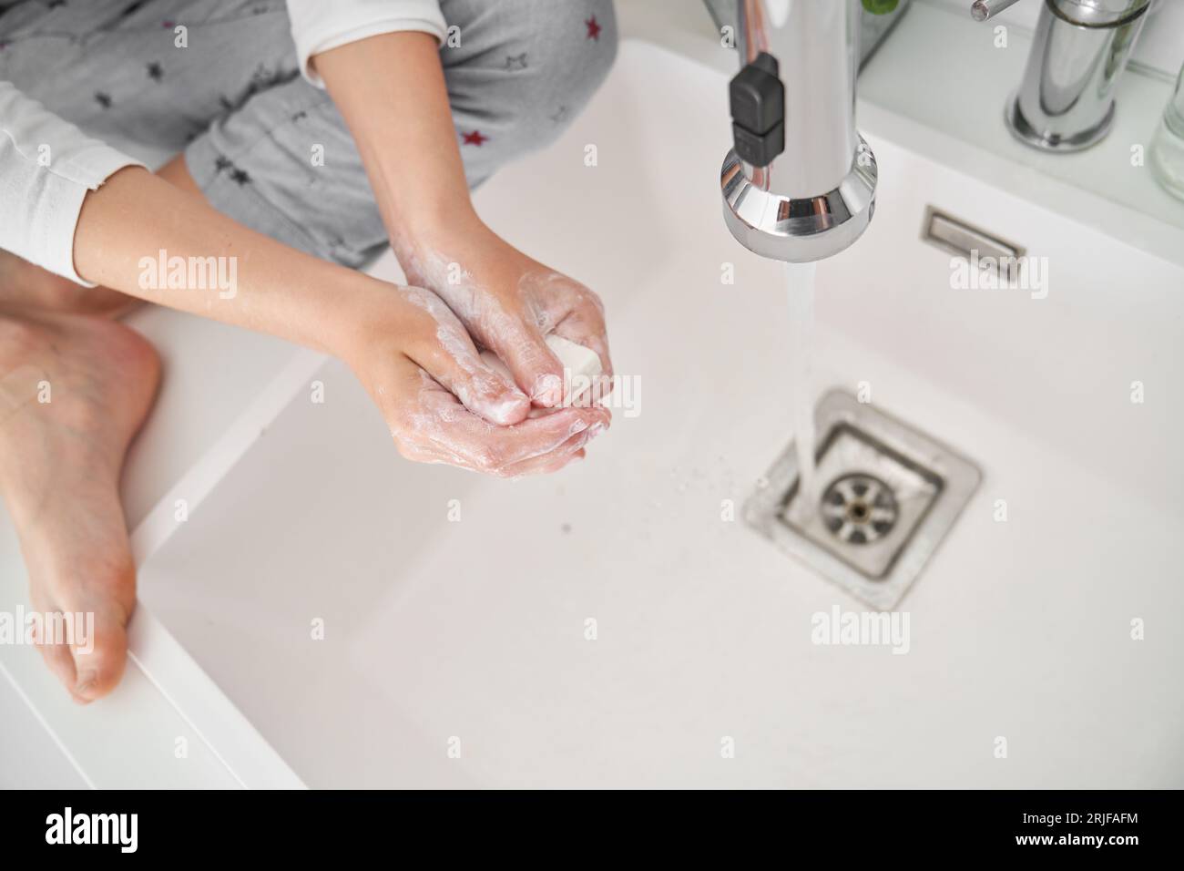 High angle of crop anonymous barefoot child sitting with crossed legs and washing hands with soap with stream of water from faucet Stock Photo