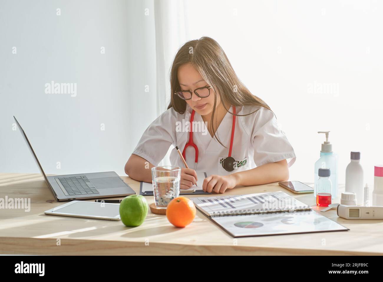 Concentrated Asian female physician in uniform taking notes on clipboard while working at table with laptop in modern light clinic Stock Photo