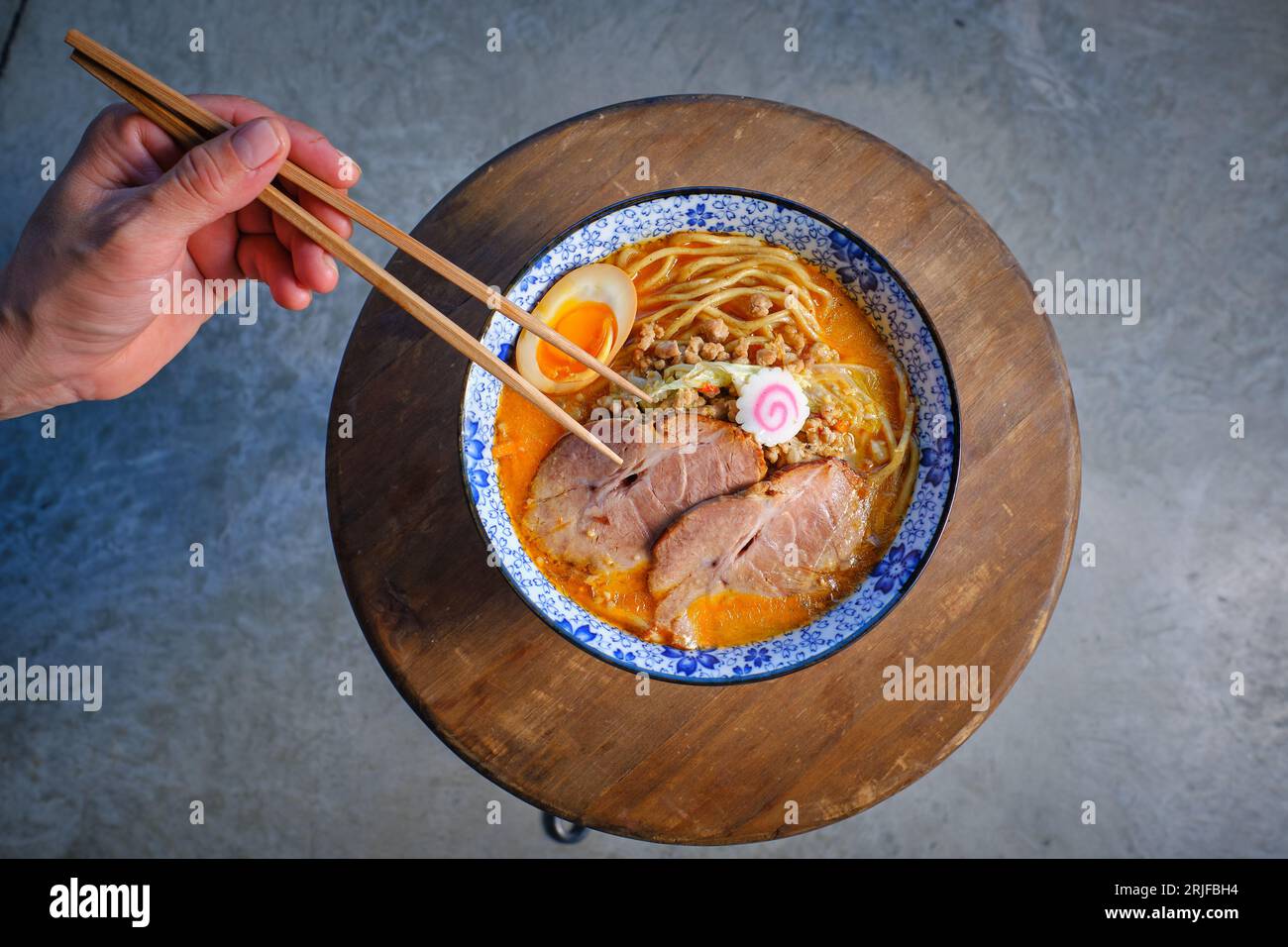 Top view of crop unrecognizable person holding chopsticks while eating delicious Japanese ramen soup with noodles and pork meat garnished with egg and Stock Photo