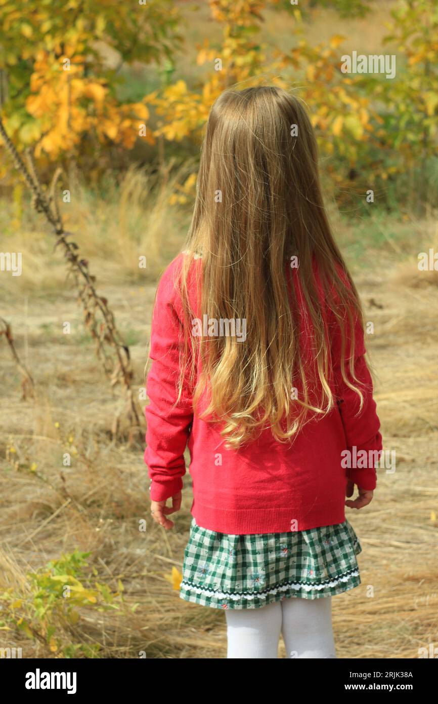 Beautiful little girl long blond hair in pink jacket plaid skirt on autumn nature background back view. Cute child walking alone in forest. Psychology Stock Photo