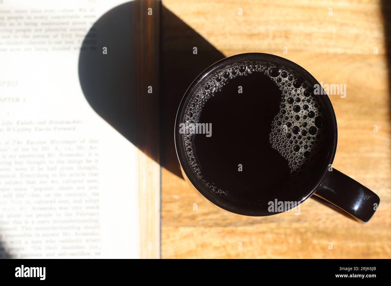 Coffee and Book. Directly above shot of black coffee and book with a coffee cup on a wood table. Image captured with bright sunlight and deep shadow. Stock Photo