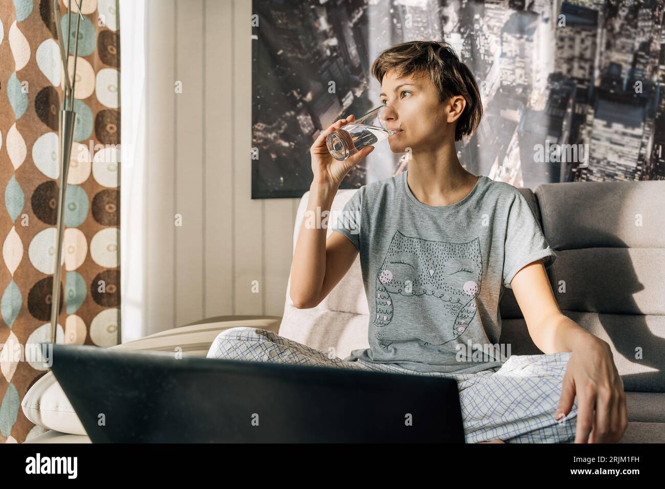 Young blonde drinks water from a glass sitting at home with a laptop. Stock Photo