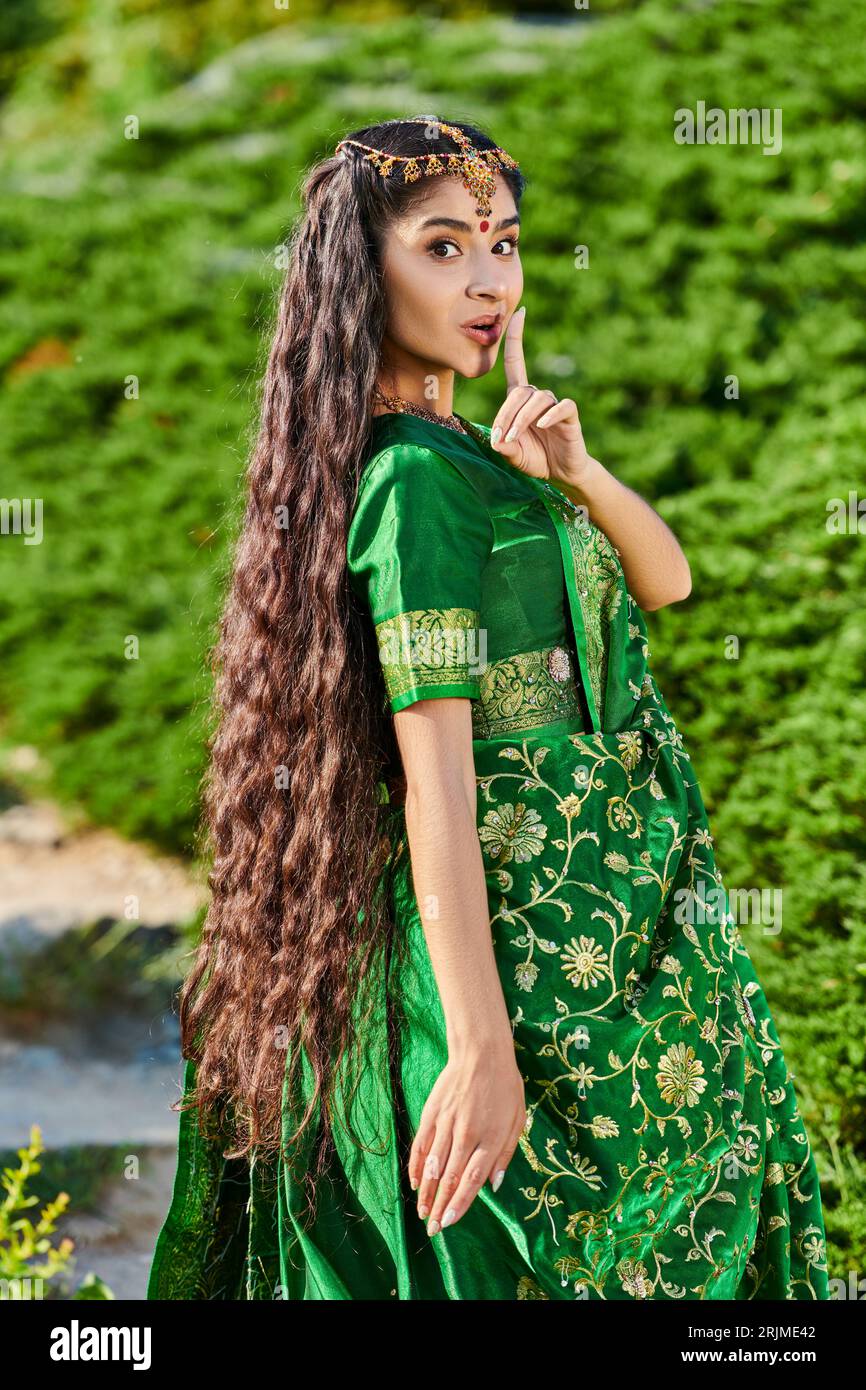 young indian woman in sari showing secret gesture and looking at camera near plants in park Stock Photo