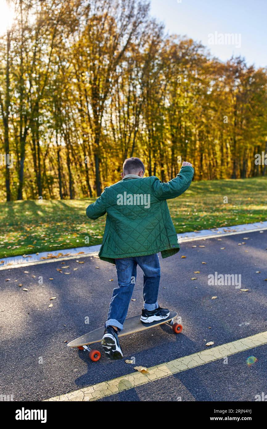 back view of boy in outerwear and jeans riding penny board in park, autumn, golden leaves, cute kid Stock Photo