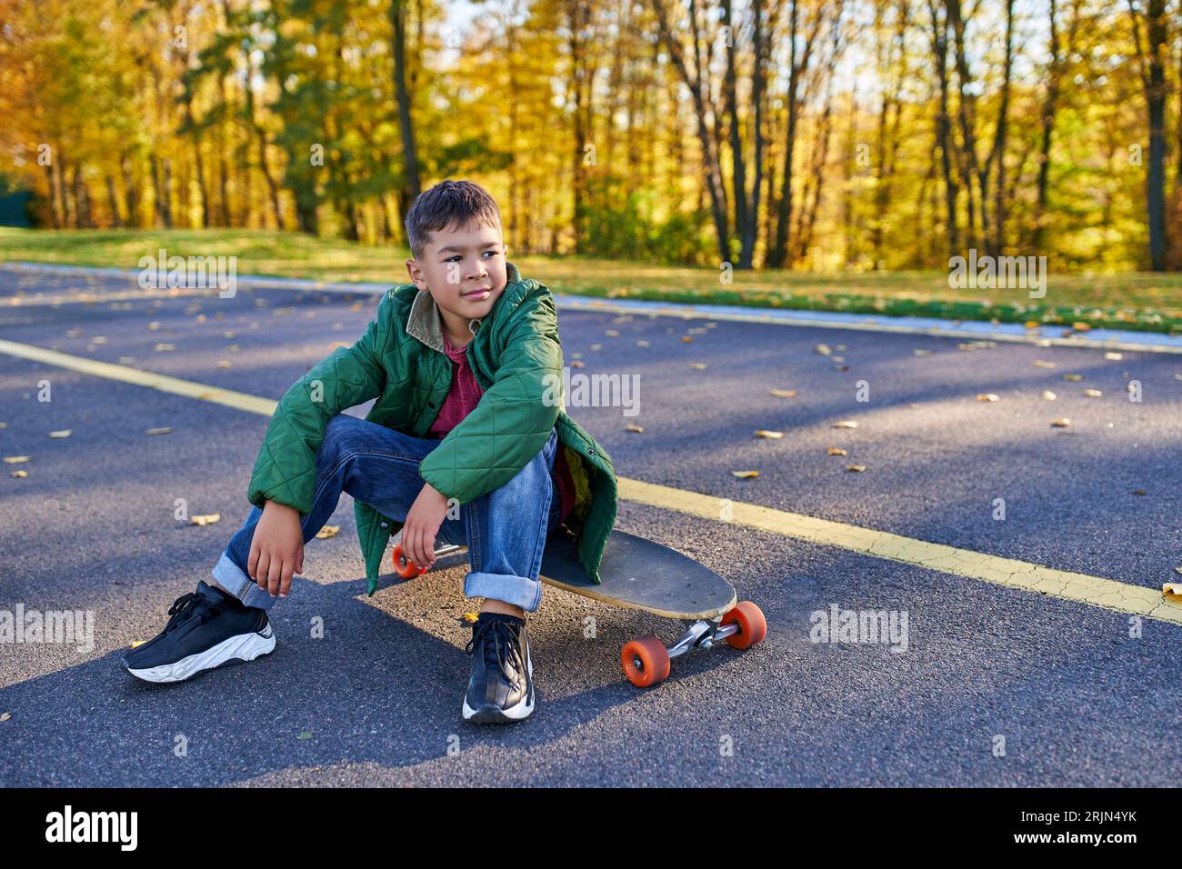pensive african american boy in outerwear sitting on penny board, autumn park, fall season Stock Photo