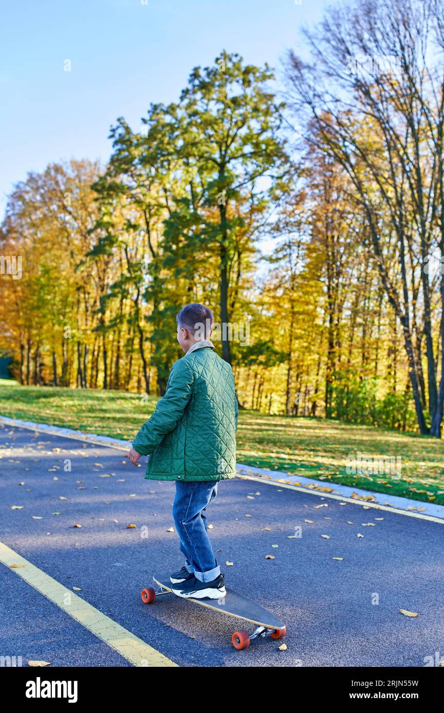boy in autumnal outerwear riding penny board, asphalt, park in fall season, golden leaves, cute kid Stock Photo