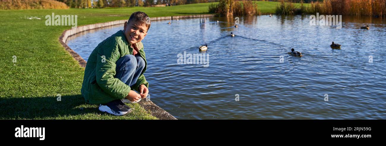 happy african american boy in outerwear sitting near pond with ducks, nature and kid, banner Stock Photo
