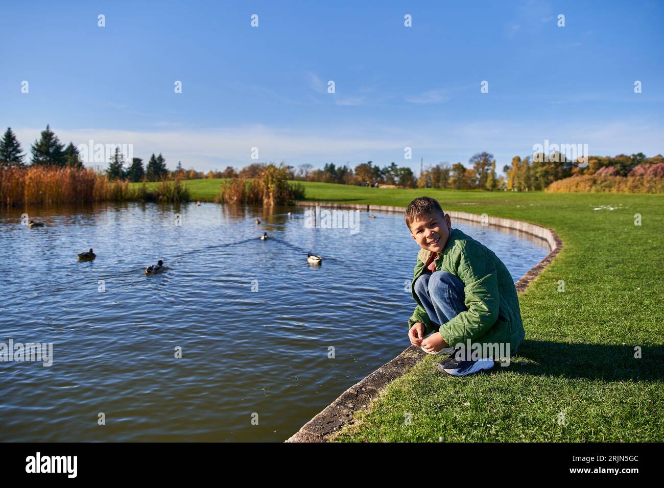 happy african american boy in outerwear and jeans sitting near pond with ducks, nature and kid Stock Photo