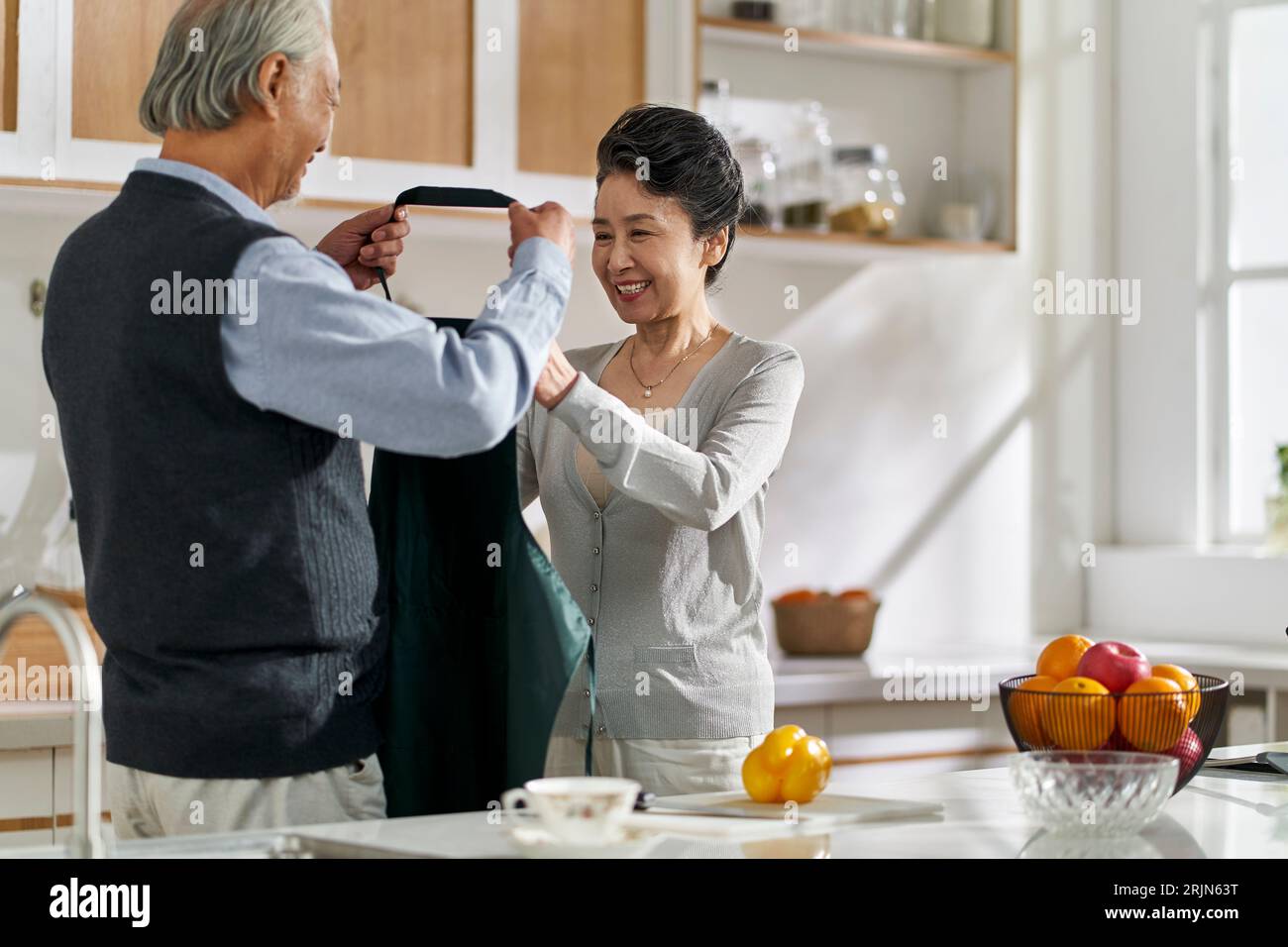 loving senior asian man husband helping wife put on apron in kitchen at home Stock Photo