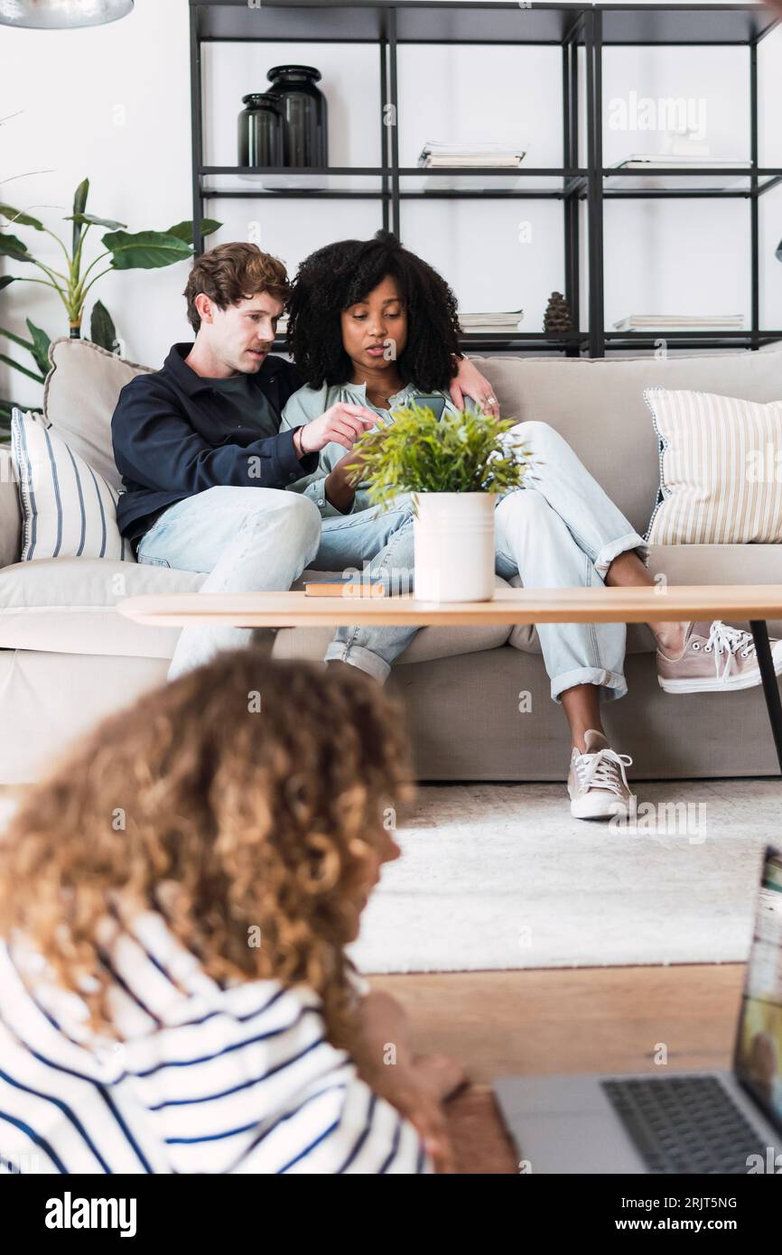 Girl having video call with friend lying on floor iwth parents sitting on couch in background Stock Photo