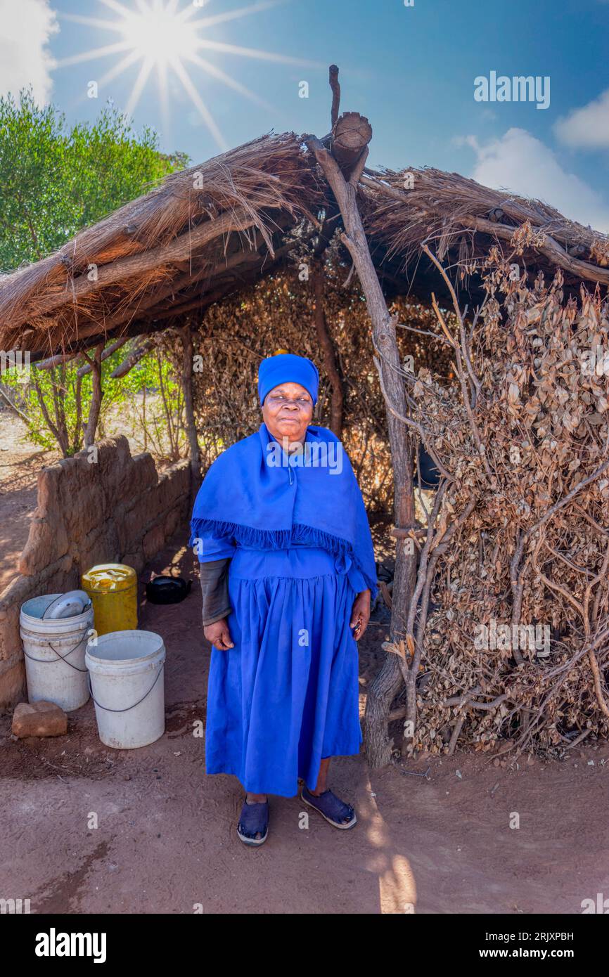 African old woman in a blue dress sited in front of her outdoors kitchen in the sandy yard, in a late afternoon day, Stock Photo