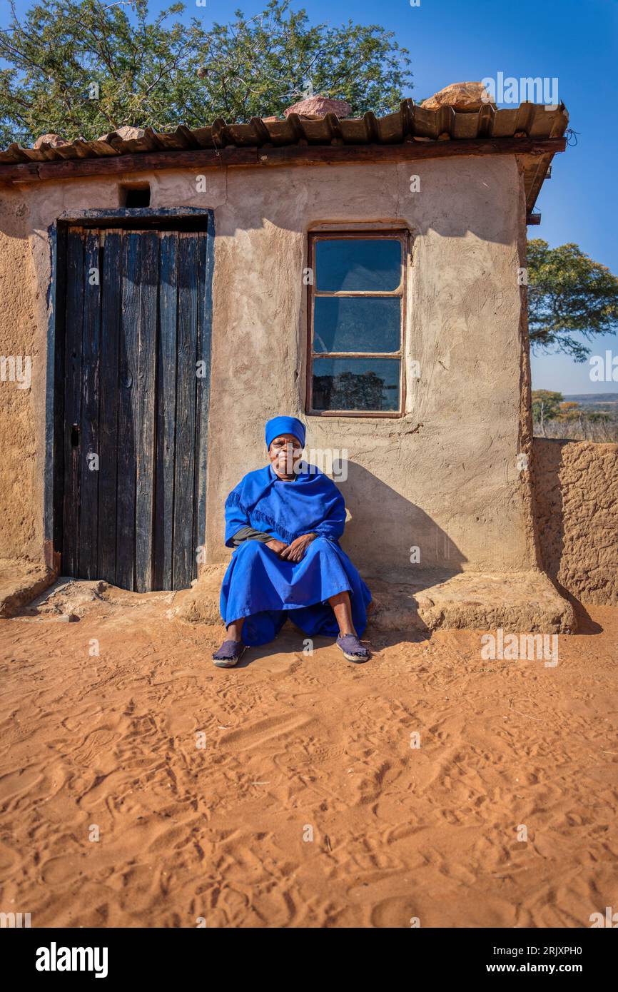 African old woman in a blue dress sited in front of her home in the sandy yard, in a late afternoon day Stock Photo