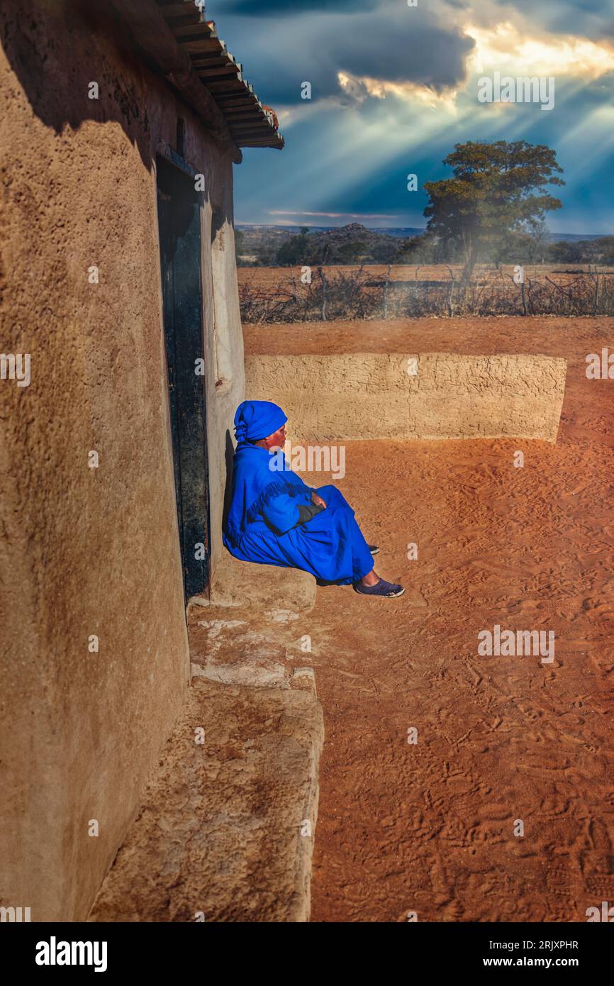 African old woman in a blue dress sited in front of her home in the sandy yard, in a late afternoon day, Stock Photo