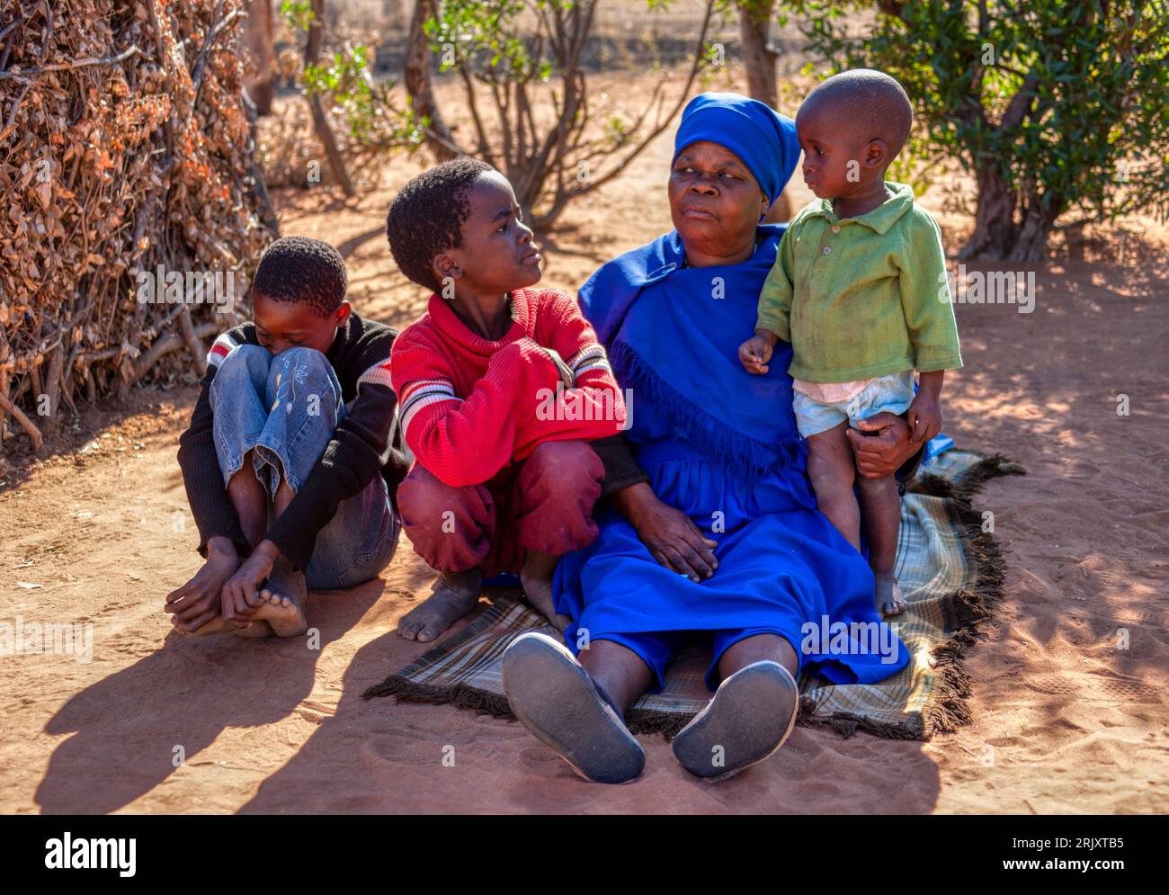 African old woman in a blue dress sited in front of her home in the sandy yard, in a late afternoon day, together with her grandchildren telling them Stock Photo