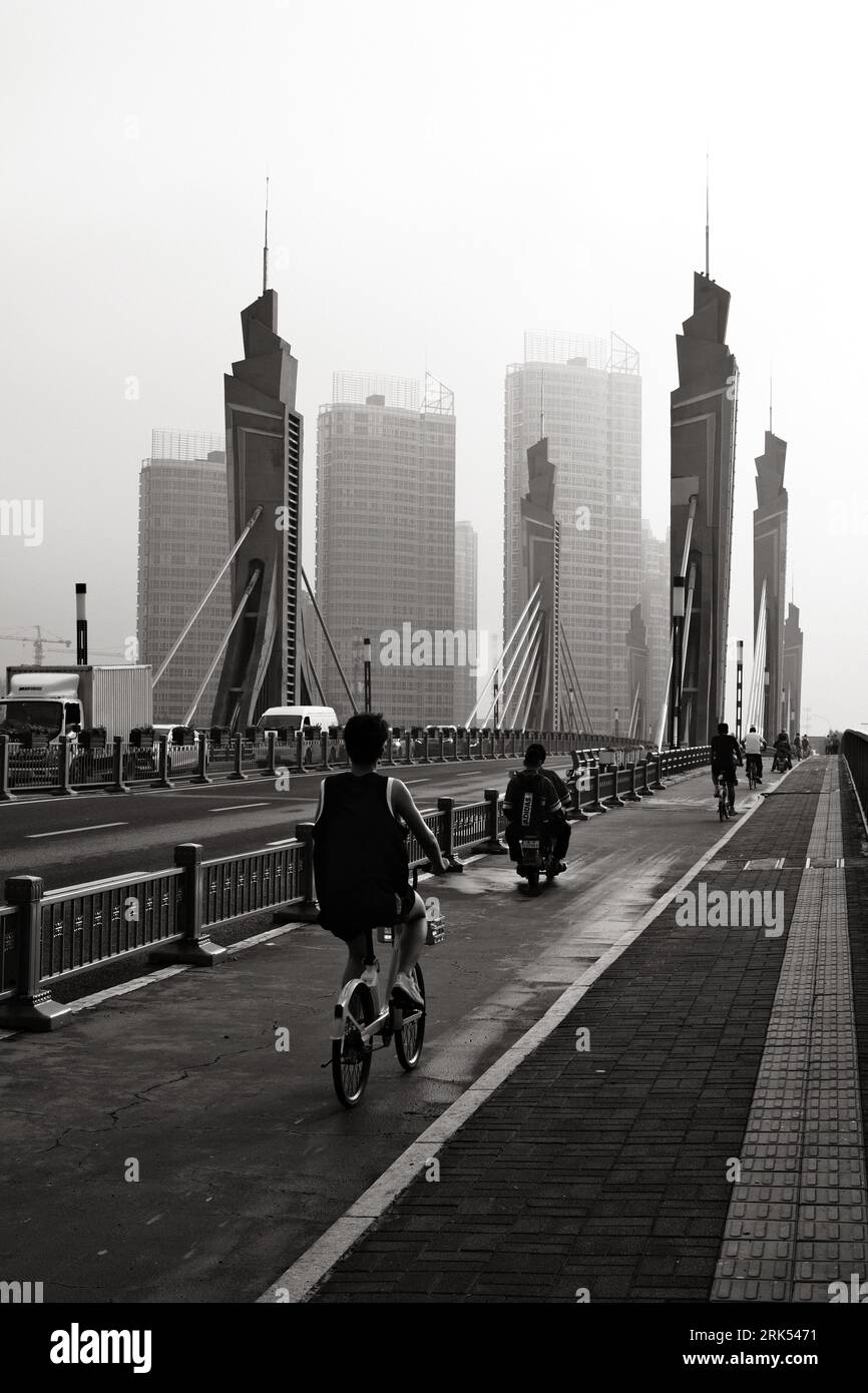 A young adult man is leisurely cycling down a bridge sidewalk, surrounded by fellow pedestrians Stock Photo