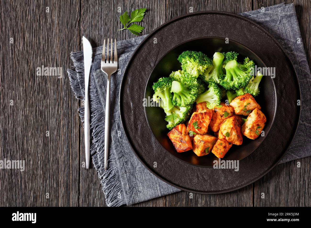 fried salmon bites with cooked broccoli florets in black bowl on dark wooden table with sriracha mayo sauce, horizontal view from above, free space Stock Photo