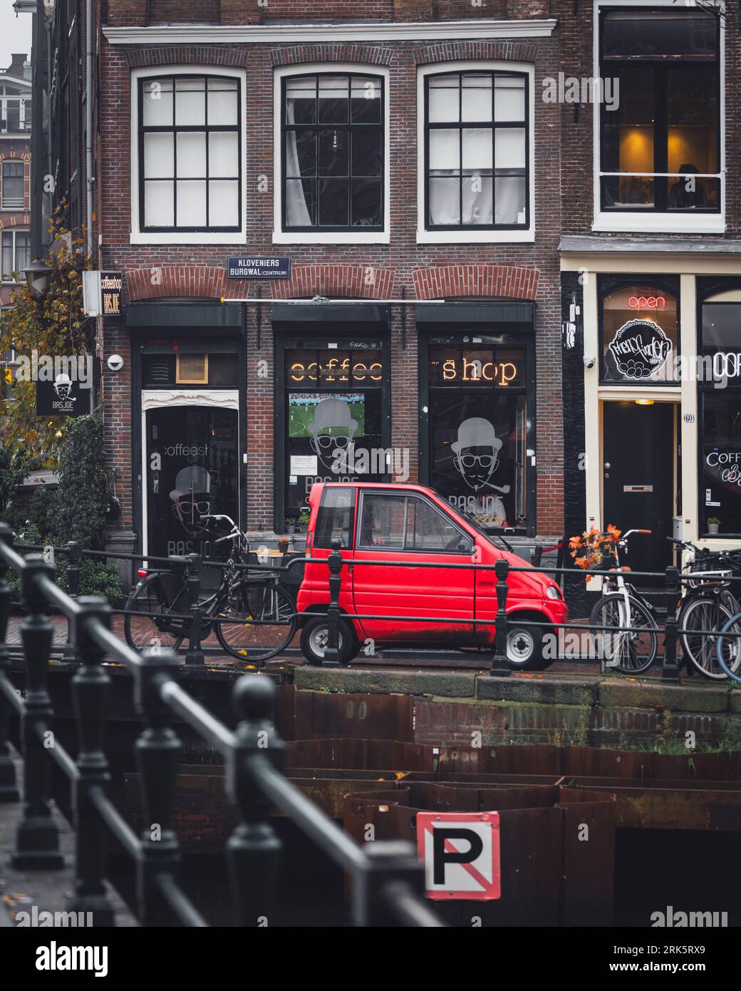 Amsterdam, Netherlands - November 27 2022: A red car is parked at the side of a canal outside a cafe and coffee shop on a street corner in Amsterdam. Stock Photo