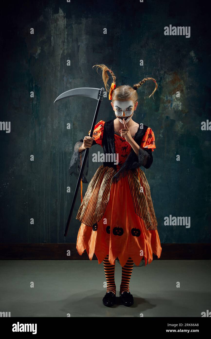 Shh. Keeping secret. Little girl with creepy makeup, costume for halloween standing with scythe against vintage green background Stock Photo