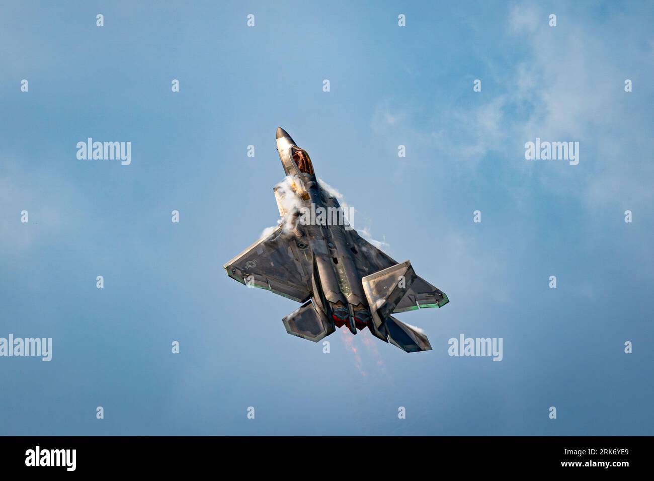 A sleek military jet fighter aircraft soaring through a bright blue sky, emitting white smoke Stock Photo