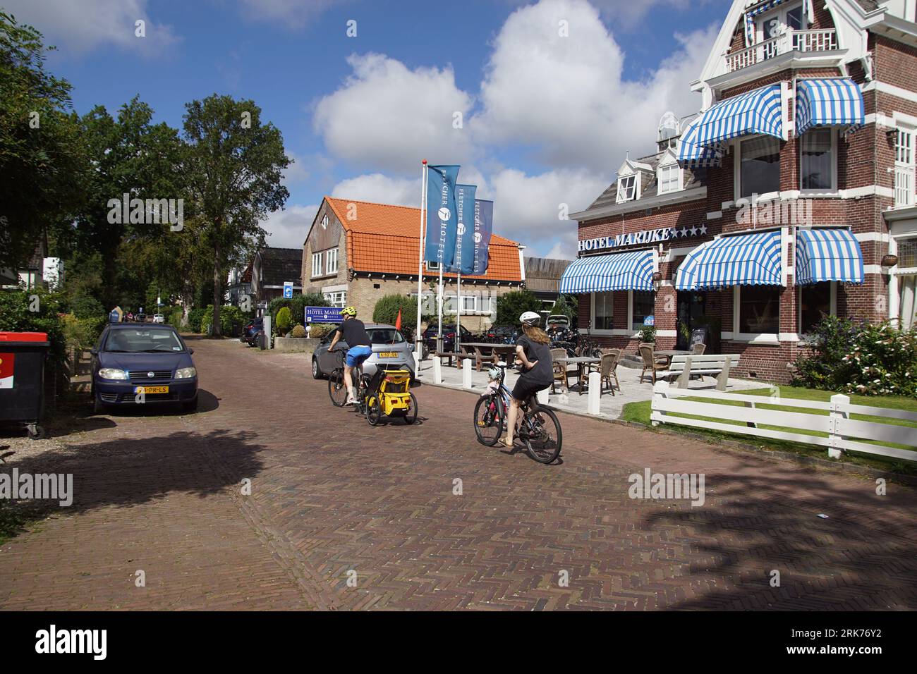 People cycle with bicycle helmets in the street called Dorpsstraat past Hotel Marijke in the Dutch village of Bergen. Netherlands, August Stock Photo